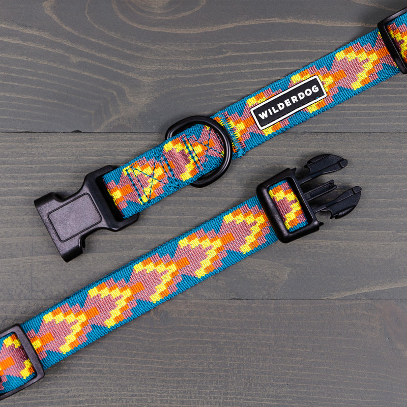 The Havasu Ultralight Collar from the Ultralight Collection is a colorful dog collar featuring a vibrant, abstract pattern in shades of blue, yellow, orange, and red. It includes a heavy-duty plastic clip and strong nylon webbing. The brand name "Wilderdog" is displayed on a fabric label. The collar is laid flat on a grey wooden surface.